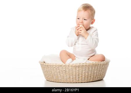 Enfant prenant l'oeuf de Pâques à la bouche en se baignant les mains, assis sur la couverture dans le panier sur fond blanc Banque D'Images