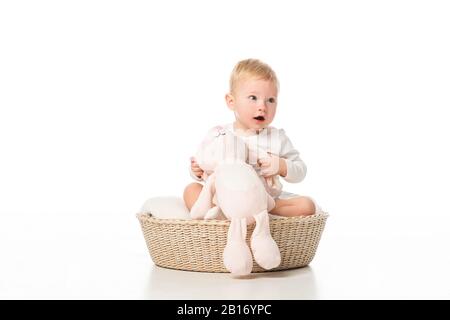 Joli enfant à col ouvert tenant un lapin rose et assis dans le panier sur fond blanc Banque D'Images