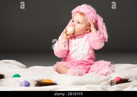 Enfant portant un costume de lapin, mettant l'oeuf de pâques dans la bouche et assis sur une couverture isolée sur le noir Banque D'Images