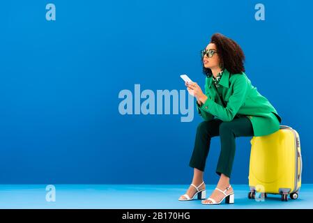 une femme africaine américaine assise sur un sac de voyage et tenant son smartphone sur fond bleu Banque D'Images