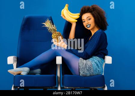un américain africain souriant assis sur des sièges et tenant des bananes et des ananas isolés sur le bleu Banque D'Images