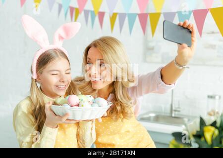 foyer sélectif de la mère heureuse et de la fille dans des oreilles de lapin prenant le selfie avec des oeufs de pâques peints dans le panier Banque D'Images