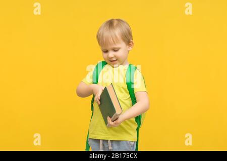 Enfant blond garçon avec un sac à dos ouvre un livre sur un fond jaune clair. Concept pour les articles sur l'étude, la lecture, l'enfance et l'enfant préscolaire Banque D'Images