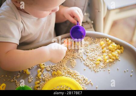 petit enfant verse une cuillère jaune jouet dans la tasse. Jeux avec produits. La méthode de développement de l'enfant de Walfdorf. Enseigner aux enfants à manger de manière indépendante. DEVE Banque D'Images