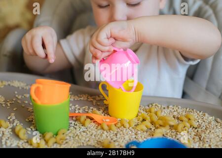 petit enfant assis sur une chaise d'alimentation et joue avec des ustensiles de jouet et du grain. développement de compétences en moteur fin chez les enfants jusqu'à l'année. Développement précoce Banque D'Images