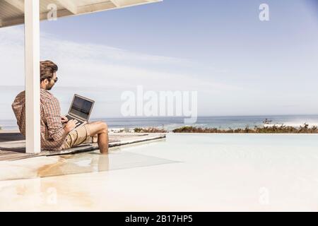 Homme travaillant sur un ordinateur portable près de la piscine dans une maison de plage Banque D'Images