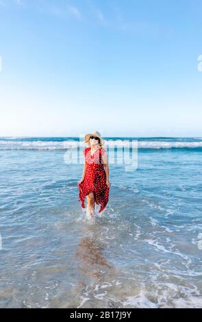 Blonde femme portant une robe rouge et chapeau à la plage, courir dans l'eau Banque D'Images