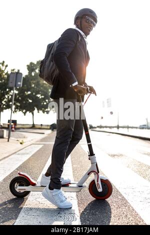 Homme d'affaires avec sac à dos et casque de vélo traversant la rue sur le scooter de poussée au coucher du soleil Banque D'Images