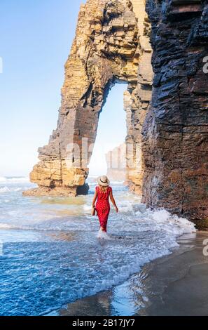 Blonde femme portant robe rouge et chapeau et marchant le long de la plage, Arche naturelle à Playa de Las Catedrales, Espagne Banque D'Images