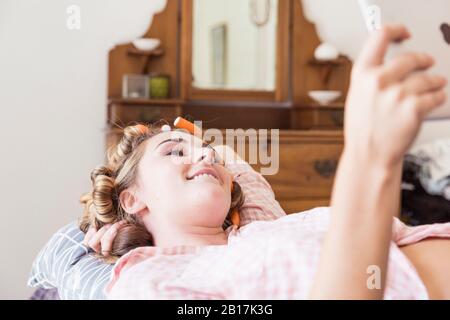 Jeune femme avec des bigoudis dans les cheveux allongé sur le lit prenant selfie avec tablette numérique Banque D'Images