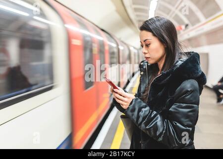 Portrait d'une femme en attente sur la plate-forme de la station de métro en regardant son smartphone, Londres, Royaume-Uni Banque D'Images