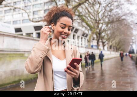 Jeune femme souriante avec téléphone portable et écouteurs dans la ville, Londres, Royaume-Uni Banque D'Images
