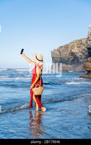 Blonde femme portant une robe et un chapeau rouges et utilisant un smartphone à la plage Banque D'Images