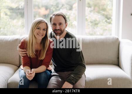 Couple assis à la maison sur le canapé, avec les bras autour Banque D'Images