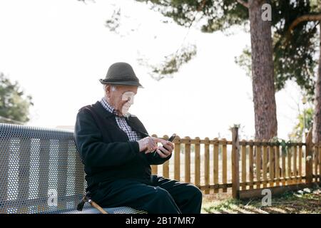 Vieil homme avec, assis sur banc, à l'aide d'un smartphone Banque D'Images