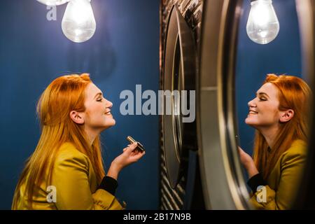 Jeune femme souriante à tête rouge regardant son image miroir appliquant le rouge à lèvres Banque D'Images