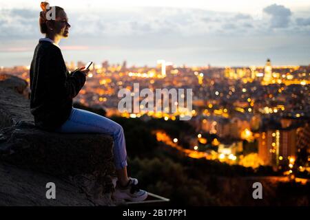 Jeune femme assise sur la rambarde au-dessus de la ville en utilisant un téléphone cellulaire, Barcelone, Espagne Banque D'Images