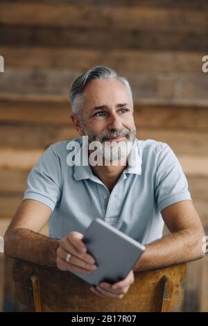 Portrait d'un homme souriant et mûr avec une tablette numérique se penchant sur le dos reposer en regardant la distance Banque D'Images