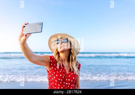 Blonde femme portant une robe et un chapeau rouges et utilisant un smartphone et prendre un selfie à la plage, faisant un embrasser la bouche Banque D'Images