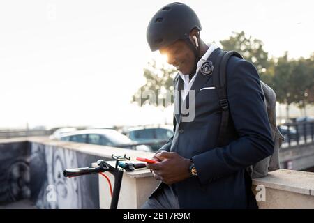Jeune homme d'affaires souriant avec scooter de poussée utilisant le téléphone mobile et les écouteurs au coucher du soleil Banque D'Images