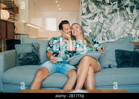 Joyeux couple assis sur le canapé dans le salon portant des chemises hawaïennes Banque D'Images