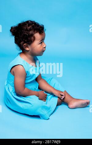 Petite fille pieds nus portant une robe bleu clair assise sur fond bleu Banque D'Images