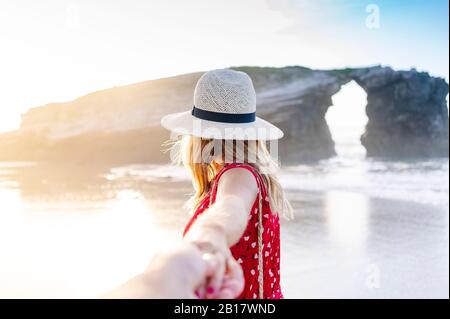 Blonde femme portant robe rouge et chapeau et tenant main sur la plage, Natural Arch à Playa de Las Catedrales, Espagne Banque D'Images