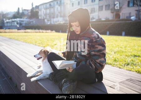 Jeune femme avec piercings et tresses assis avec son chien sur un banc de lecture d'un livre, Côme, Italie Banque D'Images