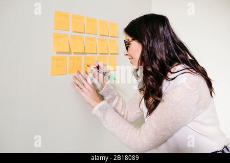 Jeune femme debout au bureau sur un mur prenant des notes sur des notes adhésives Banque D'Images