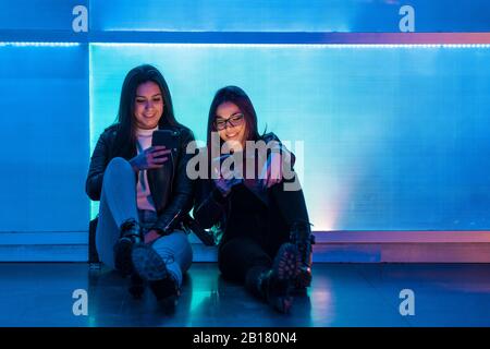 Portrait de deux adolescentes souriantes assis devant un panneau en verre bleu regardant les smartphones Banque D'Images