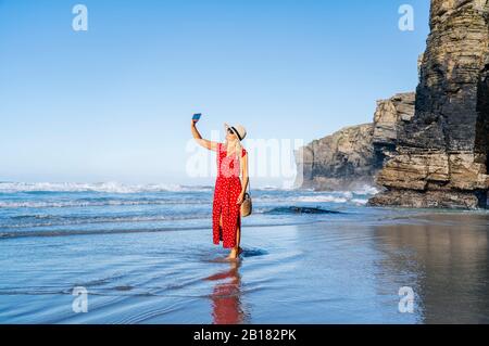 Blonde femme portant une robe et un chapeau rouges et utilisant un smartphone à la plage Banque D'Images