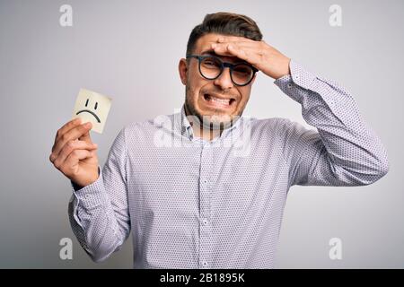 Jeune homme d'affaires aux yeux bleus tenant papier note avec un visage triste comme message malheureux souligné avec la main sur la tête, choqué avec la honte et le visage surprise Banque D'Images