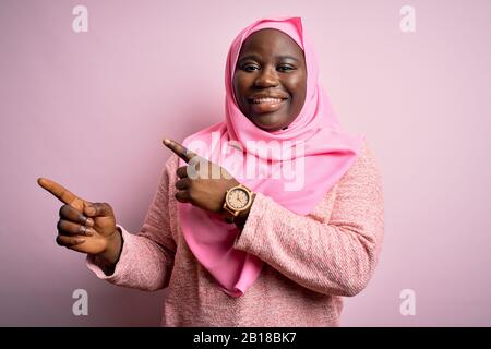 Jeune afro-américaine plus femme de taille portant un hijab musulman sur un arrière-plan rose isolé souriant et regardant la caméra pointant avec deux mains an Banque D'Images