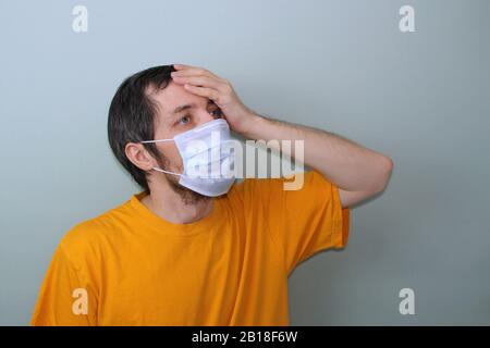 Un homme adulte d'âge moyen dans un T-shirt jaune sur fond gris dans un masque médical tient sa main près de son front. Maux de tête, forte fièvre. Problème d'épidémie de coronavirus de protection contre les maladies en provenance de Chine. Banque D'Images