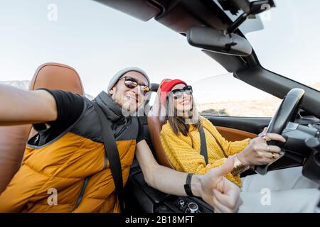 Portrait selfie d'un couple heureux en conduisant une voiture de sport convertible sur la route du désert pendant un coucher de soleil. Style de vie et concept de voyage insouciant Banque D'Images