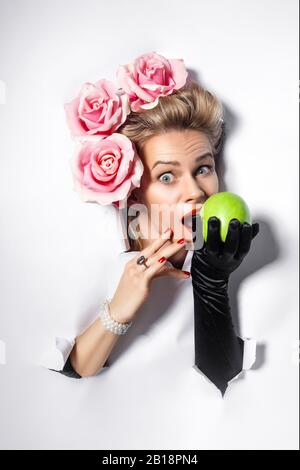 une femme gaie fait un trou dans le livre blanc. Une femme regarde à travers un trou dans un fond blanc de papier. Une femme morde une pomme. Une femme décorée Banque D'Images
