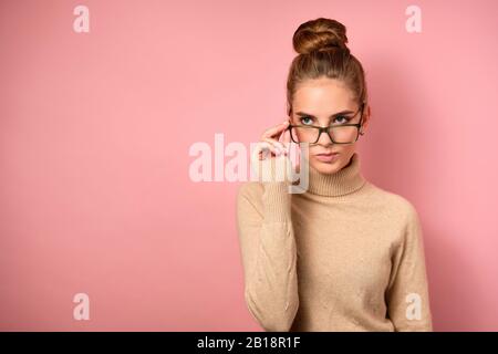 Une fille avec une peau propre et un haut pain se tient sur le fond rose et semble décontent du côté au-dessus des verres. Banque D'Images