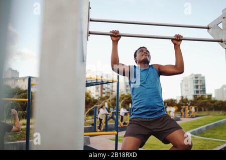 Vue de face d'un athlète masculin africain fort faisant monter sur le bar horizontal dans le parc public de la salle de sport Banque D'Images