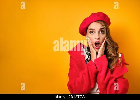 Une fille dans un manteau rouge et beret regarde en choc sur le cadre, l'ouverture de sa bouche et la pression de ses mains sur son visage. Banque D'Images