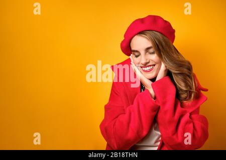 Une fille dans un manteau rouge et un béret est debout sur un fond jaune et gentille calmant et souriant, appuie ses paumes sur son visage Banque D'Images