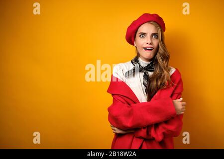 Une belle fille dans un manteau rouge et un béret, avec un mouchoir de cou se tient sur un fond jaune avec ses yeux larges et la bouche tordue Banque D'Images