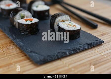 Des petits pains à sushis au saumon, au thon, à l'avocat, au riz aux algues et des baguettes sur un tableau sombre allongé sur un tapis de bambou. Banque D'Images