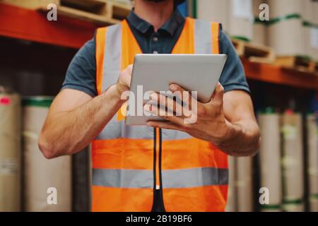 Gros plan du travailleur de l'entrepôt dans la veste orange qui maintient la tablette numérique en état de marche tout en vérifiant l'inventaire et la commande Banque D'Images