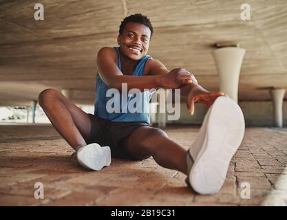 Vue de face d'un jeune homme souriant portant des baskets qui s'étirent sur sa jambe avant de courir en ville Banque D'Images