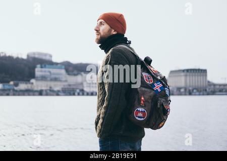 Jeune voyageur barbu avec des patchs sur le sac à dos debout au bord de la rivière avec paysage urbain sur fond. Banque D'Images