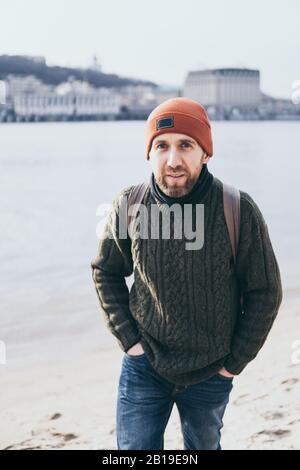 Jeune voyageur barbu avec sac à dos debout au bord de la rivière avec paysage urbain sur fond. Regarder dans l'appareil photo. Banque D'Images