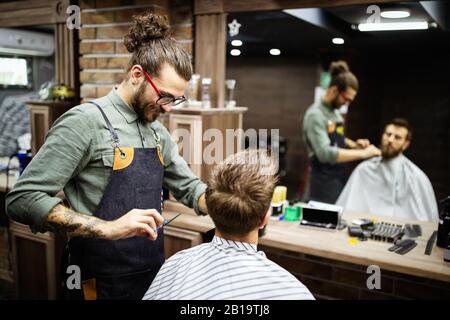 Jeune homme barbu se coupe par coiffure while sitting in chair at barbershop Banque D'Images