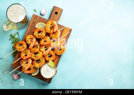 Brochettes de crevettes grillées ou langoustines servies avec de la chaux, de l'ail et de la sauce sur fond de béton bleu clair. Fruits de mer et bière. Vue de dessus avec copie Banque D'Images