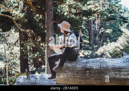 Une femme aux cheveux rouges avec un chapeau et un sac à dos, en traversant une forêt en regardant une carte assise sur un tronc d'arbre Banque D'Images