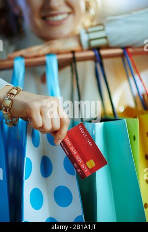 Gros plan sur heureuse élégante femme de 40 ans en pull blanc et jupe avec carte de crédit près des sacs de shopping colorés accrochés sur des vêtements en cuivre rail. Banque D'Images